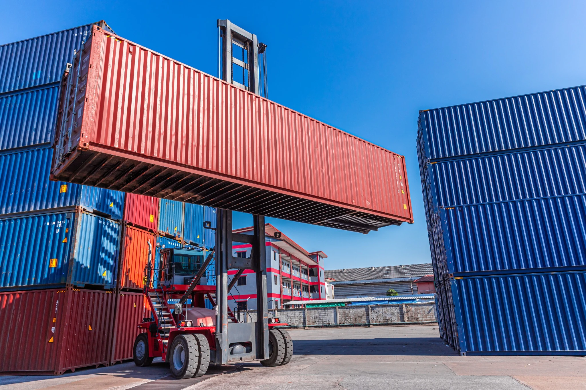 A heavy-duty container handler lifts a red cargo container at a shipping terminal in Ontario, Canada.